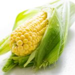 Detailed close-up of a fresh ear of corn with water droplets, showcasing vibrant yellow kernels and green husks.